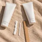 Two plain white unbranded sunscreen tubes on a sand-coloured beach towel in strong sun, with one thick line and one thin line of white cream squeezed beside a wooden ruler